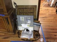 Photo showing a group of framed artworks leaning against a wooden cabinet and wall on a hardwood floor, including a large family-themed metal sign, bathroom art print, and beach landscape.