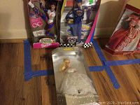 Four vintage Barbie dolls in original boxes: Wedding Barbie, NASCAR Barbie, Olympic Gymnast Barbie, and a fourth Barbie in a red dress, arranged on a wooden floor with blue tape markings.
