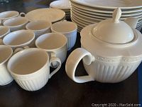 Photo of 12 white cups, white teapot with lid, and stacks of plates and saucers showing embossed pattern