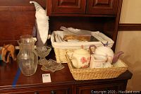Lot includes decorative ceramic tea pots, crystal candy dishes, woven baskets, and small decorative items arranged on a wooden surface with wood-paneled backdrop.
