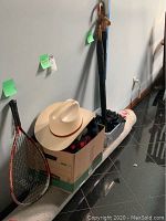 Photo showing the white cowboy-style hat sitting on top of a box containing scarves and gloves, with a tennis racket leaning against the box and two umbrellas standing nearby.