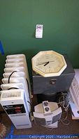 Photo showing multiple white electric space heaters stacked, an octagonal wall clock, an electric ballast, a desk phone, and white paper trays all placed against a green wall.