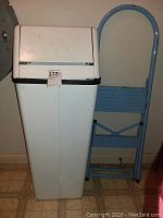 Photo showing blue metal step ladder and white plastic waste basket with flip lid side by side.
