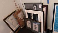 Five pieces of artwork and frames standing on floor against a wall showing floral prints and black and white portraits.