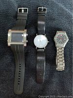 Three watches laid on dark fabric: Arnette digital with large rectangular face and black rubber strap, Levenger analog with clean white face and black leather strap, Wenger analog with black dial and metal bracelet.