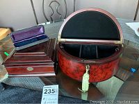 Three wooden boxes displayed on a glass table, including one semi-circular jewelry box with a tassel and lock, and two rectangular watch boxes, one with purple velvet interior.