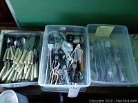 Photo showing three plastic bins containing cream-handled flatware, assorted kitchen utensils, and additional flatware items.