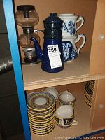 Shelf view showing glass vacuum coffee brewer, blue enamel coffee pot, white floral mugs and decorative cups with saucers