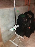 Metal adjustable art easel shown alongside stacked black luggage and bags against a basement wall.