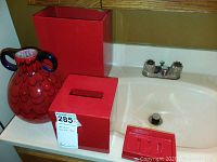 Photo showing a large decorative red art glass vase with black handles beside a red tissue box cover and a red soap dish on a bathroom sink.