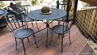 Photo showing the full outdoor table and three chairs from an angled side view on a wooden deck, set with a decorative hat on the table.