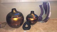 Three items on a tabletop: two bronze-colored round vases and two abstract sculptures, one black and the other purple and beige.