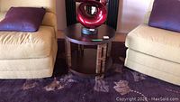 Wood round side table placed between two beige chairs on a patterned rug, showing top of table and decorative red sculpture on top.