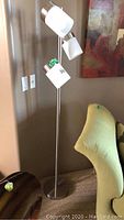 Full view of the floor lamp showing the three frosted glass shades and brushed silver pole standing next to a green chair.