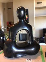 Front view of abstract black polished statue showing hollow circular cutout in torso area and smooth human sitting figure shape.