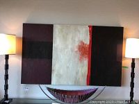 Front view of full abstract canvas painting hung on wall between two lamps with decorative bowl below.