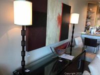 Pair of matching metal floor lamps with white drum shades placed behind a glass console table in a living room, showing overall size and style.