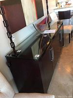 Side view showing the full length and height of the dark wood side buffet with glass top and smooth finish. Four cabinet doors visible with metal handles.