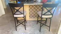 Two black metal barstools with beige microfiber seats, positioned at a kitchen counter. Chairs show signs of wear including spots and scuffs.
