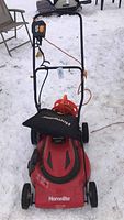 Homelite electric lawn mower standing on snow-covered ground with extension cord wrapped and attached at the handle.