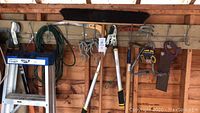 Full view of ladder with assorted hanging gardening tools including shears, hand rake, saw, and broom, along with extension cords and hose on left