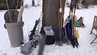 Photo showing large grey garbage can with lid, flexible hose, wire mesh rolls, watering can, garden soil bags, and folding cart near tree in snow.