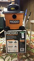 Front view of assembled Ridgid wet/dry vacuum on top of its original box. Orange tank with black top and wheels at base. Brand name Ridgid clearly visible.