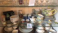 Wide view of Asian ceramic kitchen lot including teapot, cups, bowls, and other items on glass shelf.