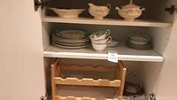 China cabinet shelf holding stack of plates, teacups with saucers, and wooden wine rack beneath