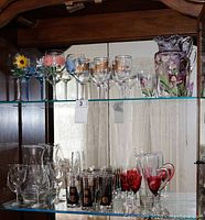 Shelf with assorted handpainted and etched glassware including wine glasses with floral designs in yellow, pink, blue, and gold tones; red glasses; clear tumblers; water pitchers; and martini mixer.