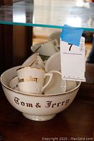 Photo showing bowl and several mugs stacked inside it, all handpainted with Tom & Jerry lettering in gold and black, with a glass shelf above.