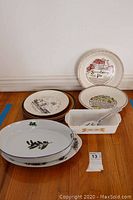 Wide view of various ceramic plates and a rectangular casserole dish with pie cutter shown on floor with a lot number card