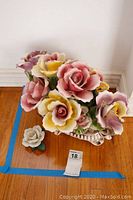Porcelain Capodimonte floral centerpiece with large pink, yellow, purple flowers and ornate base, placed on wood floor next to a small separate porcelain flower