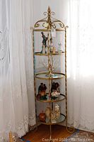 Metal rack with four round glass shelves, standing near window with lace curtains, items displayed on shelves