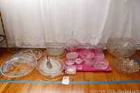 Wide shot showing entire collection of clear glass and crystal bowls, plates, platters and punch bowl positioned on wood floor with white curtain background.