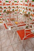 Full view of the rectangular wrought iron table with clear glass top, showing the 4 matching chairs with orange cushions on tiled floor and patterned wallpaper.