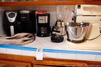 Photo of kitchen countertop showing five kitchen appliances: Power Air Fryer XL (black), Keurig coffee maker (black, no pot), Better Chef coffee maker (black/stainless steel), Oster blender (stainless steel base, glass jar), and Sunbeam Mixmaster stand mixer (cream color) with some wear visible.