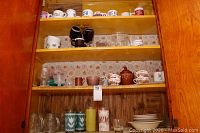 Upper shelves showing coffee mugs with floral and decorative designs, black and brown mugs, glass tumblers, and patterned teacups with saucers.
