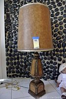 Front view of large decorative table lamp with brown shade and ornate carved wooden base against a black pebble wall background.