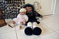 Three dolls displayed on tile floor: Jesmar baby doll in pink dress and bonnet, large rag doll in green dress and hat, and background cloth doll with printed face.