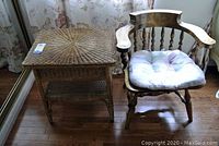 Wicker side table and wooden armchair with cushion shown side by side on wooden floor near floral curtains.