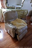 Front view of light beige leather recliner rocker chair with crochet throw showing padded armrest and cushioned back.