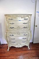 Front view of a tall boy dresser with cream finish, ornate floral carvings on the drawer fronts, and decorative metal handles.