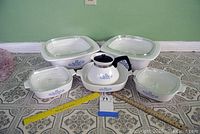 Full view of six CorningWare bakeware pieces, white with Blue Cornflower pattern and clear lids, arranged on patterned tiled floor with rulers for scale.