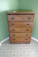 Front view of vintage 5-drawer wooden dresser showing wear on top and drawer fronts, brass-tone handles, and curved front edge on top surface.