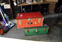 Two foot lockers stacked, red on top and green below, showing exterior wear and brass hardware