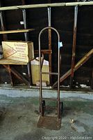 Metal hand truck with rust on the frame, standing upright in a garage environment with wooden shelves in the background.