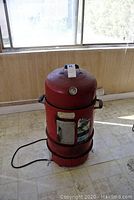 Red electric smoker shown from the front with temperature gauge, door, and electrical cord visible. Positioned on a tiled floor near a window.