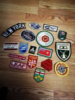 Top-down view of 17 assorted vintage cloth patches displayed on wood floor with various regional and thematic designs from Canada, New York and safety emblems.
