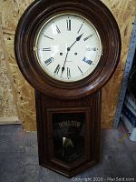 Full front view of wooden regulator clock showing round dial bezel and lower pendulum door